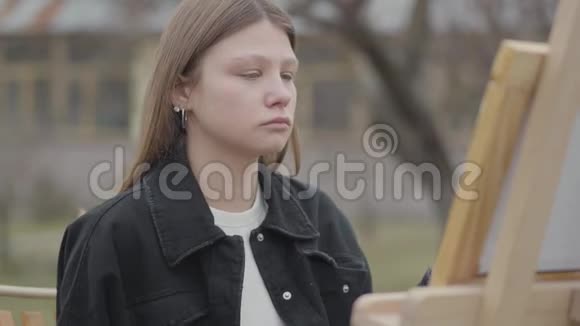 坐在木制画架前的年轻女画家画一幅画抽烟休闲女艺术家视频的预览图