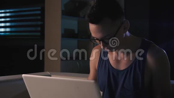 一个戴眼镜的年轻人在黑暗的房间里的笔记本电脑后面工作视频的预览图