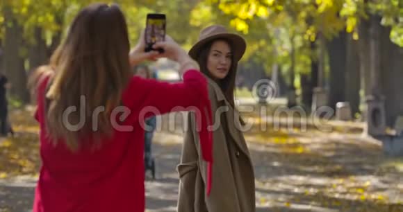 美丽的高加索女人穿着优雅的棕色外套和帽子在秋天公园的背景下为她的女朋友摆姿势视频的预览图