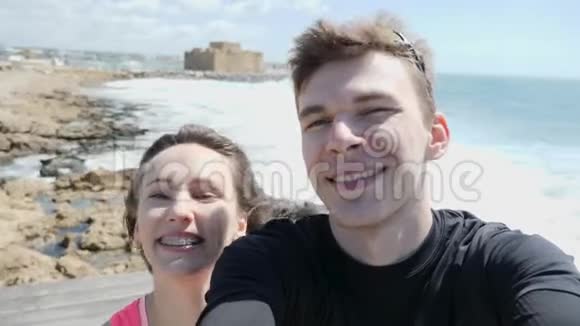 年轻快乐的情侣在岩石海滩上以自拍模式微笑着进入镜头强烈的波浪撞击岩石视频的预览图