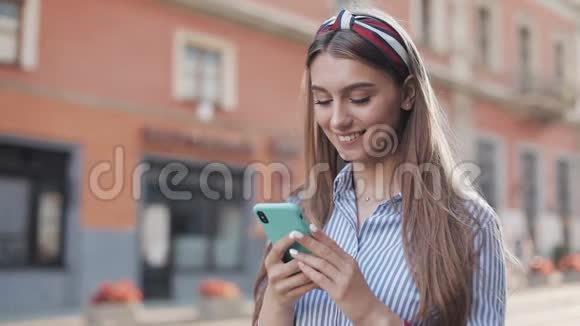 年轻时尚迷人的白人女孩棕色头发头带着她的智能手机滚动和微笑视频的预览图