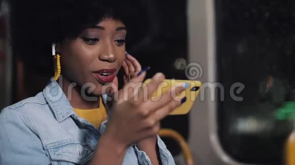 微笑着年轻的非洲裔美国妇女握着扶手在公共交通工具上看智能手机上的视频夜晚视频的预览图