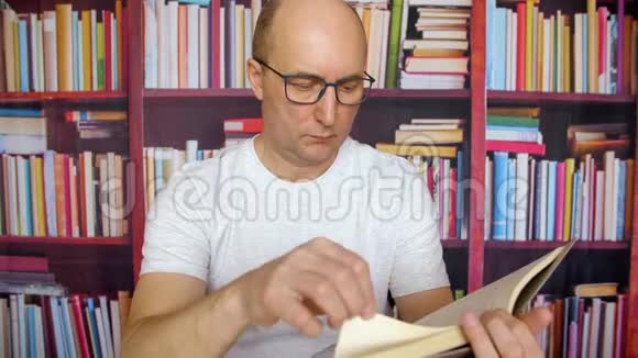 体贴的白人在图书馆内部的书架背景下读书戴着眼镜和白色t恤的男人坐在视频的预览图
