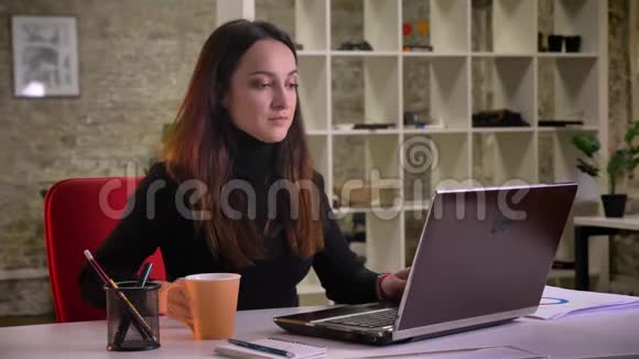 年轻的黑发女商人在笔记本电脑上打字和在办公室喝咖啡的肖像视频的预览图