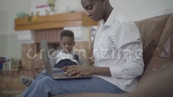 一位在笔记本电脑上工作的非裔美国妇女和她可爱的小儿子在舒适的客厅里玩玩具视频的预览图