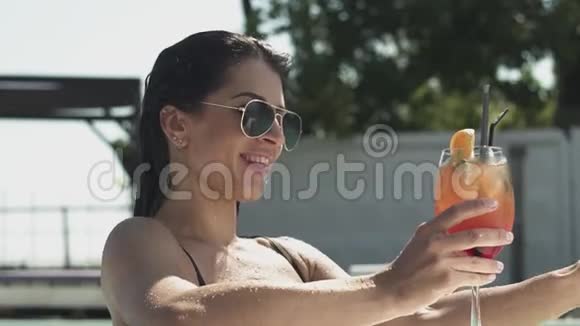 戴太阳镜的漂亮女人在游泳池里摆着鸡尾酒的肖像穿比基尼的孤独女士的休闲视频的预览图