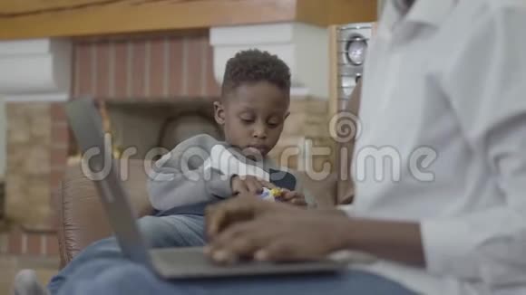 可爱的非裔美国妇女在笔记本电脑上工作她的小可爱儿子在舒适的客厅附近玩玩具视频的预览图