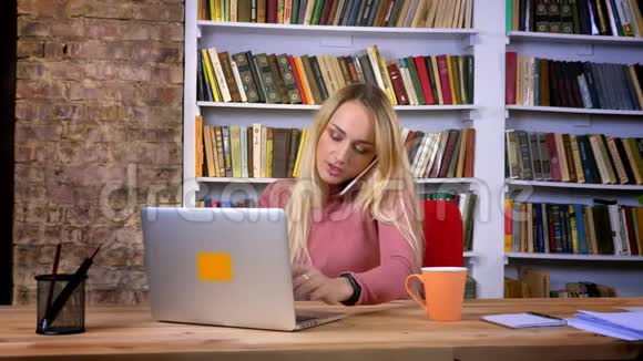 年轻的白种人女孩的肖像她穿着漂亮的衣服专心地用笔记本电脑工作在书架上用手机说话视频的预览图