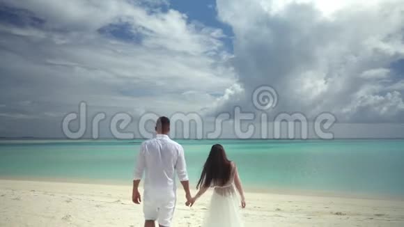 新婚夫妇手牵手在一片绚丽的白色沙滩和绿松石水上背景中是一片美丽的天空热带婚礼视频的预览图