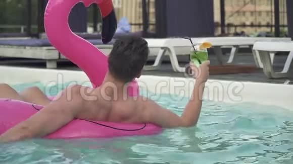 年轻的滑稽男子在游泳池边游到充气的火烈鸟旁边游泳的是性感的黑发和可爱的金发女郎视频的预览图