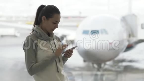 年轻女子使用智能手机站在机场窗口附近背景是飞机视频的预览图