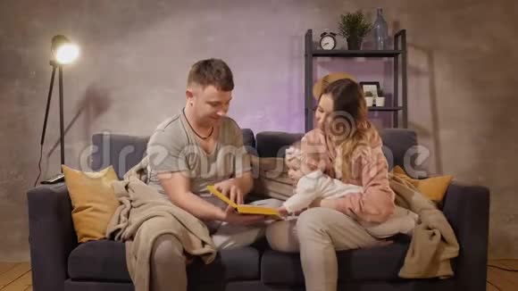 一个年轻的家庭坐在舒适的房间里的沙发上妈妈爸爸和宝宝在看书家庭幸福视频的预览图