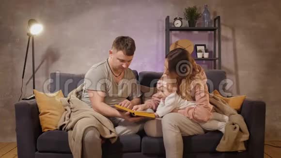 一个年轻的家庭坐在舒适的房间里的沙发上妈妈爸爸和宝宝在看书家庭幸福视频的预览图
