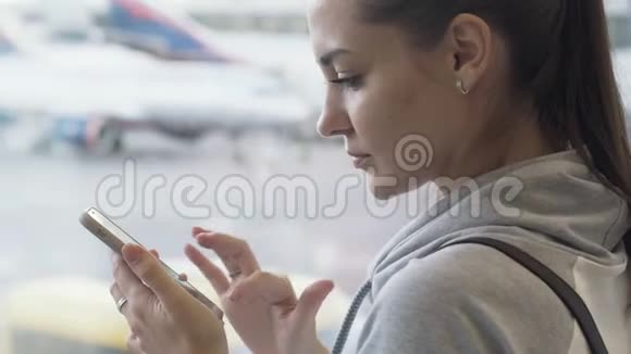 侧视吸引女孩使用智能手机和短信在机场背景的飞机窗口特写视频的预览图