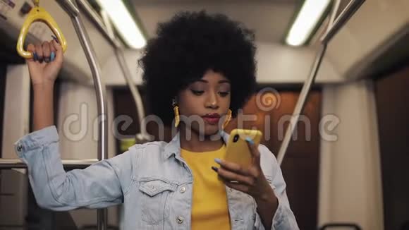 漂亮的非裔美国年轻女性握着扶手在公共交通中使用智能手机晚上关闭视频的预览图