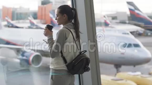 运动服装中的侧视女孩在机场背景下使用电话和饮料外卖咖啡在窗口视频的预览图