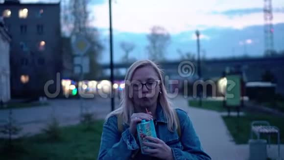 一个穿着牛仔夹克戴着眼镜的女孩在晚上的长廊上用管子喝果汁计划不错视频的预览图