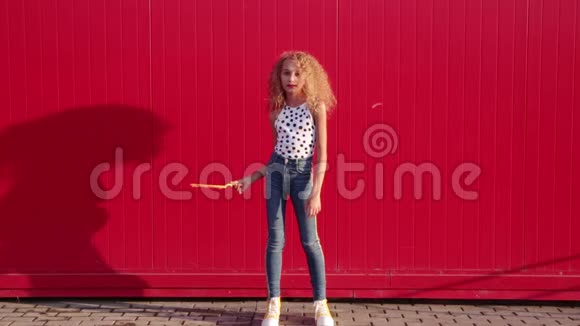 穿着豌豆印花背心和蓝色牛仔裤的卷曲金发女孩对着红墙吹肥皂泡视频的预览图
