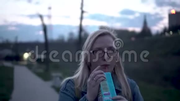 女孩沿着河边的晚堤散步通过管子从一个盒子里喝果汁视频的预览图