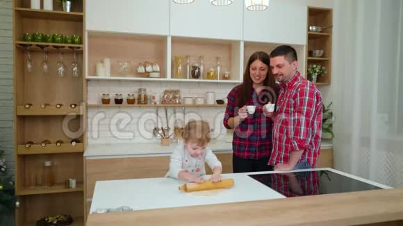 可爱的小女孩在她快乐的父母在厨房里喝咖啡的时候卷面团视频的预览图