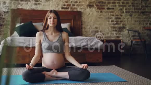 怀孕的年轻女人坐在瑜伽垫上的莲花姿势手掌放在膝盖上呼吸和放松健康视频的预览图
