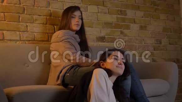 年轻的白种人女孩坐在地板上躺在朋友的膝盖上在舒适的家庭氛围中看电影视频的预览图