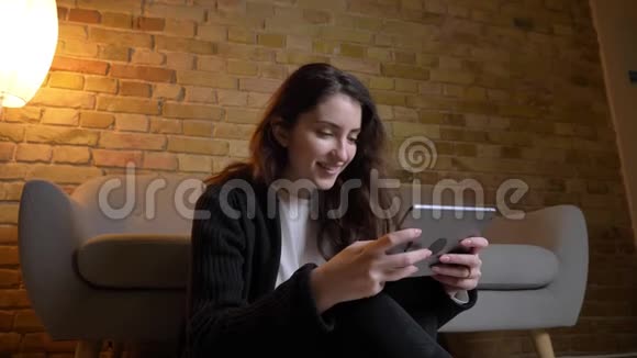 白种人女孩的肖像一头黑发坐在地板上微笑着在舒适的家里看着平板电脑视频的预览图