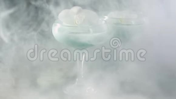 玻璃酒杯蓝色鸡尾酒和白色兰花笼罩在烟雾中视频的预览图