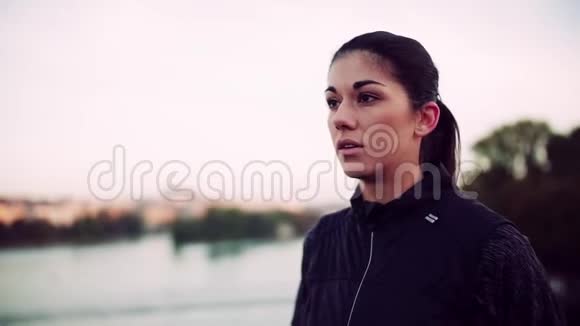 一位年轻的女跑步者站在城市的桥上戴上耳机视频的预览图