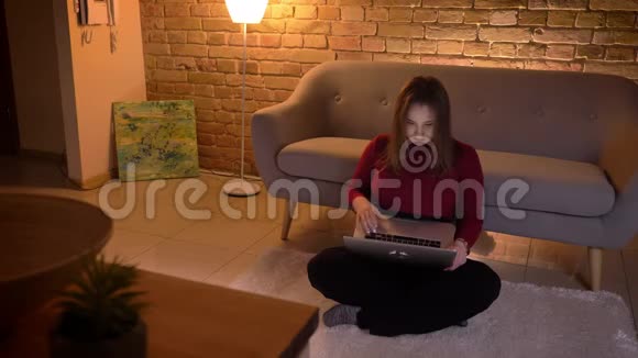 年轻漂亮的白种人女性坐在室内地板上浏览笔记本电脑的特写镜头视频的预览图