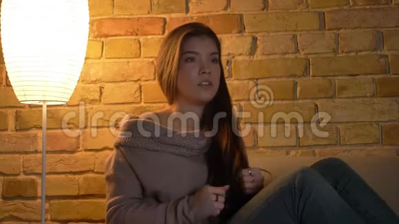 一幅被吓坏的年轻白种人女孩坐在沙发上在舒适的家庭背景下看电视的特写肖像视频的预览图