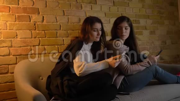 年轻的白种人女孩坐在沙发上在舒适的家庭氛围中愉快地观看他们的智能手机的肖像视频的预览图