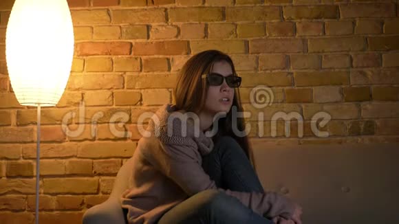 在舒适的家里戴着3D眼镜坐在沙发上看电视恐怖片的年轻白人女孩的画像视频的预览图