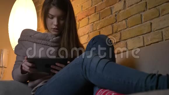 从底部的年轻白种人女孩坐在沙发上观看平板电脑在舒适的家庭背景下视频的预览图