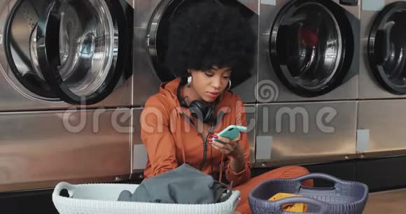 非裔美国年轻女性使用智能手机在洗衣前用篮子分拣衣服她坐着视频的预览图