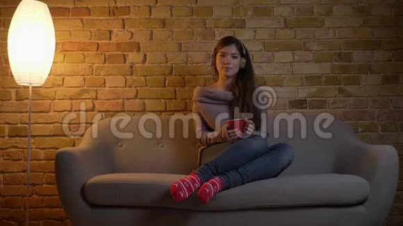 年轻美丽的白种人女孩戴着耳机坐在沙发上带着红色的杯子在布里肯墙背景上跳舞视频的预览图