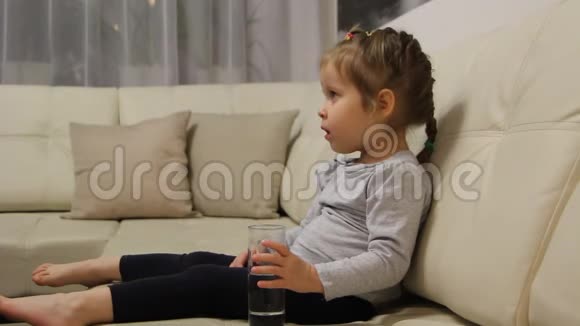 小孩子坐在房间的沙发上喝着玻璃杯里的水视频的预览图