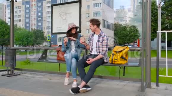 年轻夫妇在户外喝咖啡坐在公共汽车站的长椅上视频的预览图