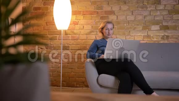 高加索大龄妇女坐在沙发上用笔记本电脑打字的特写镜头视频的预览图