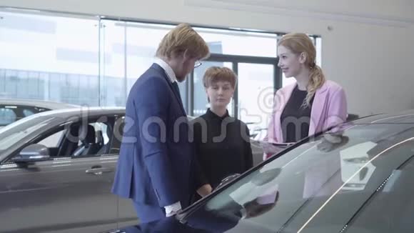 西装革履的大胡子推销员告诉两个成功的年轻女孩关于一家汽车经销店的一辆新车的信息视频的预览图