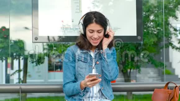 年轻女子戴着耳机欣赏音乐坐在现代有轨电车车站等公共交通视频的预览图