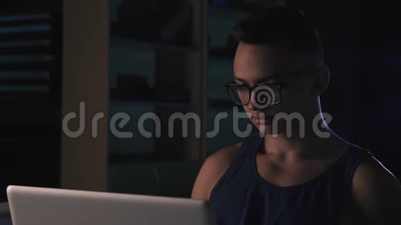 一个戴眼镜的年轻人在黑暗的房间里的笔记本电脑后面工作视频的预览图