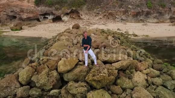 一个穿着白色裤子和衬衫的人站在靠近海洋的石头上的鸟瞰图视频的预览图