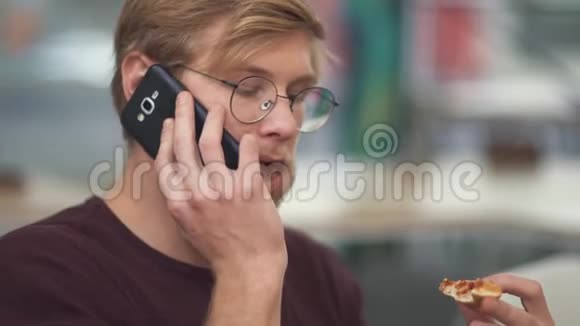 一个戴眼镜吃三文治用手机说话的留胡子的年轻人的画像视频的预览图