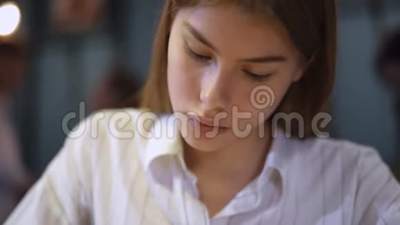 一个穿着白色衬衫的年轻女孩坐在靠近嘴唇的咖啡馆里拿着一支钢笔的肖像女孩写字然后看视频的预览图