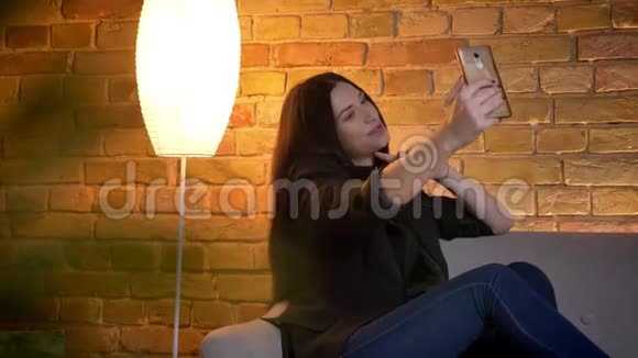 年轻的黑发白种人女孩坐在沙发上在家庭氛围中制作自拍照片的肖像视频的预览图
