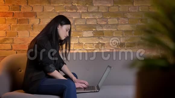 年轻的黑发白种人女孩在家庭氛围中坐在沙发上用笔记本电脑专心打字视频的预览图
