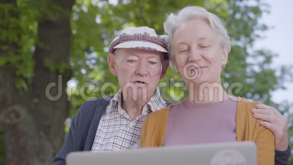 一位白发老人和一位戴帽子的老人坐在美丽的公园里的长凳上用笔记本电脑和孩子们聊天视频的预览图