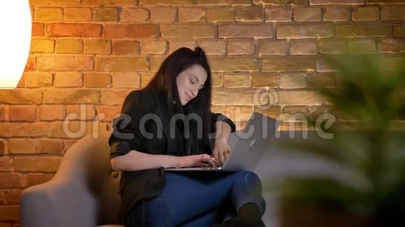 肖像画中年轻的黑发白种人女孩在沙发上观看笔记本电脑满意的家庭氛围视频的预览图