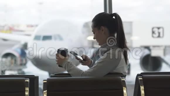 美丽的女孩喝外卖咖啡在机场候机室用电话发信息视频的预览图